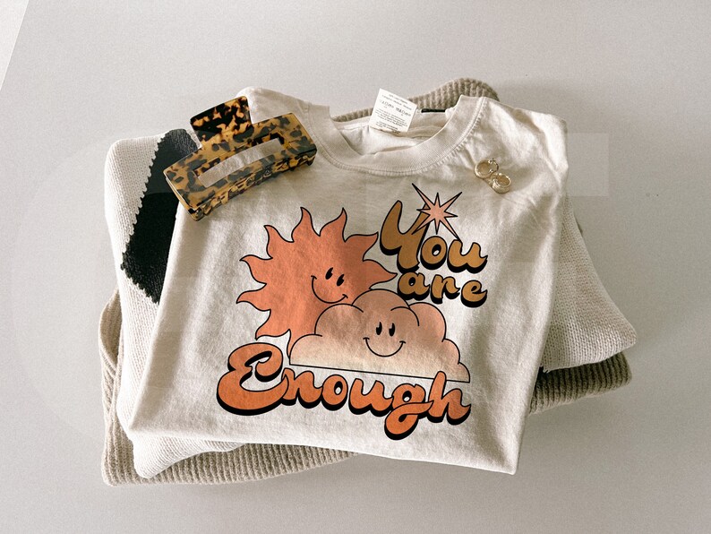 Puede incluir: Una camiseta color crema con el texto "You are Enough" en naranja y marr&oacute;n, con un sol y una nube sonrientes. Una pinza para el pelo carey y pendientes dorados descansan sobre un su&eacute;ter doblado.