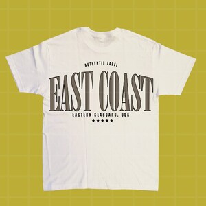 Może przedstawiać: Biała koszulka z napisem "EAST COAST" dużymi, ciemnoszarymi literami. Nad tekstem znajduje się "AUTHENTIC LABEL", a pod nim "EASTERN SEABOARD, USA" z pięcioma gwiazdkami. Koszulka wykonana jest z miękkiego materiału.