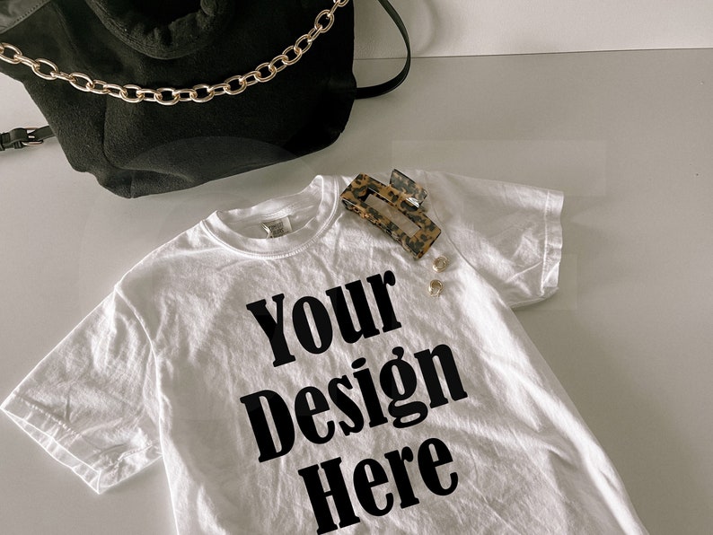 May include: White t-shirt with the text "Your Design Here" in black. A black bag with a gold chain strap is in the background. A leopard print hair clip and two gold earrings are on the shirt.