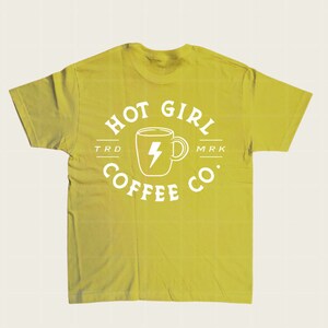 Puede incluir: Una camiseta verde amarillenta con un gr&aacute;fico blanco. El gr&aacute;fico presenta una taza de caf&eacute; con un rayo en el interior y las palabras "HOT GIRL COFFEE CO." en un dise&ntilde;o circular. La camiseta tiene cuello redondo y mangas cortas.