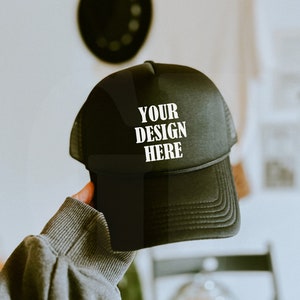 Może przedstawiać: Czarna czapka typu trucker z białym napisem "YOUR DESIGN HERE". Czapka ma siatkowy tył i zakrzywiony daszek. Czapka jest trzymana przez osobę.