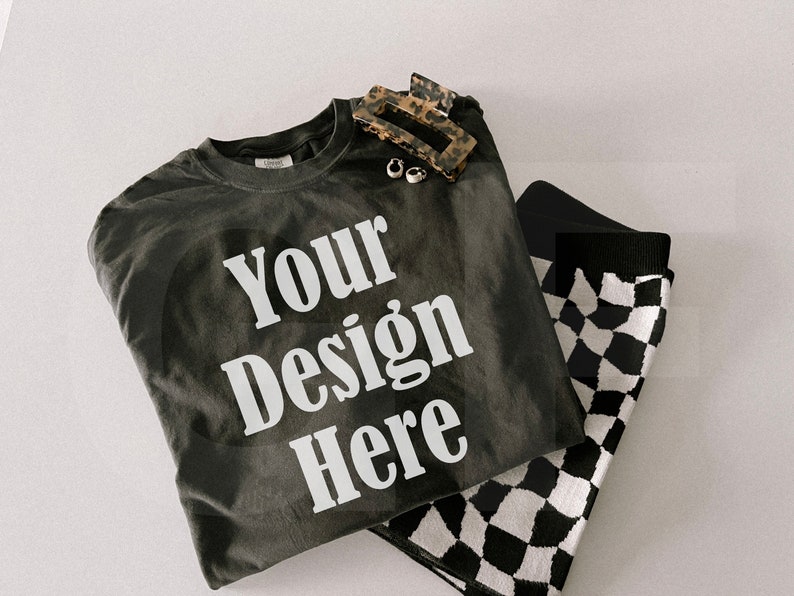May include: A dark gray t-shirt with the text "Your Design Here" in white, paired with black and white checkered socks, a tortoiseshell hair clip, and small silver earrings. The items are arranged on a white surface.