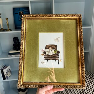 May include: A framed watercolor illustration featuring a brown armchair with a small dog sitting on it, a side table with flowers, and a floor lamp. The artwork is encased in a textured gold frame with a wide, olive green velvet matting border.