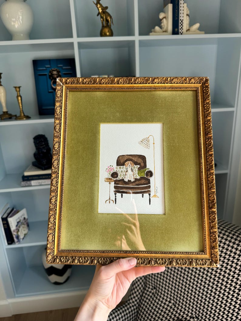 May include: A framed watercolor illustration featuring a brown armchair with a small dog sitting on it, a side table with flowers, and a floor lamp. The artwork is encased in a textured gold frame with a wide, olive green velvet matting border.
