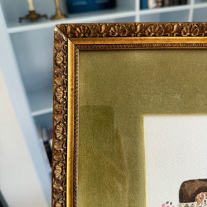 May include: A framed artwork featuring an ornate, antique-style gold frame with a textured, scalloped edge. The frame includes a wide, olive green velvet mat surrounding a white paper print that depicts a dog sitting near a small floral arrangement.