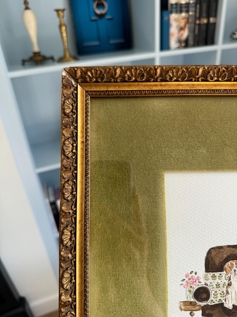 May include: A framed artwork featuring an ornate, antique-style gold frame with a textured, scalloped edge. The frame includes a wide, olive green velvet mat surrounding a white paper print that depicts a dog sitting near a small floral arrangement.