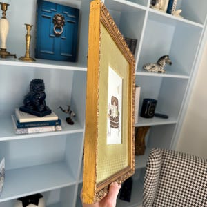 May include: A rectangular picture frame with an ornate gold finish and a textured green matting. The frame contains a small illustration of a brown armchair with a white dog sitting on it, accompanied by the text "BE KIND TO YOURSELF".
