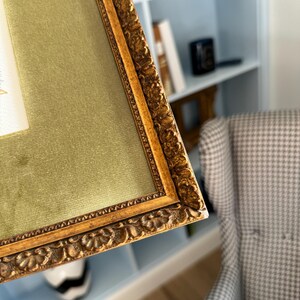 May include: A rectangular picture frame featuring an ornate, textured gold-toned border with intricate floral-like carvings. The frame includes a wide, smooth olive green velvet matting that surrounds a central white rectangular space.