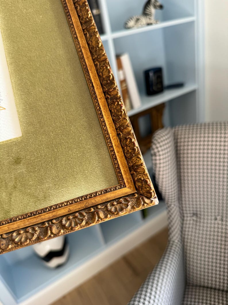 May include: A rectangular picture frame featuring an ornate, textured gold-toned border with intricate floral-like carvings. The frame includes a wide, smooth olive green velvet matting that surrounds a central white rectangular space.