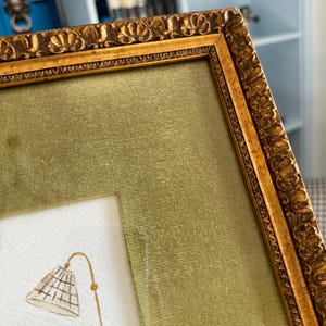 May include: A rectangular picture frame with an ornate, textured gold finish and a wide, olive green velvet mat. Inside the frame, a white paper surface features a watercolor illustration of a floor lamp with a grid-patterned lampshade.