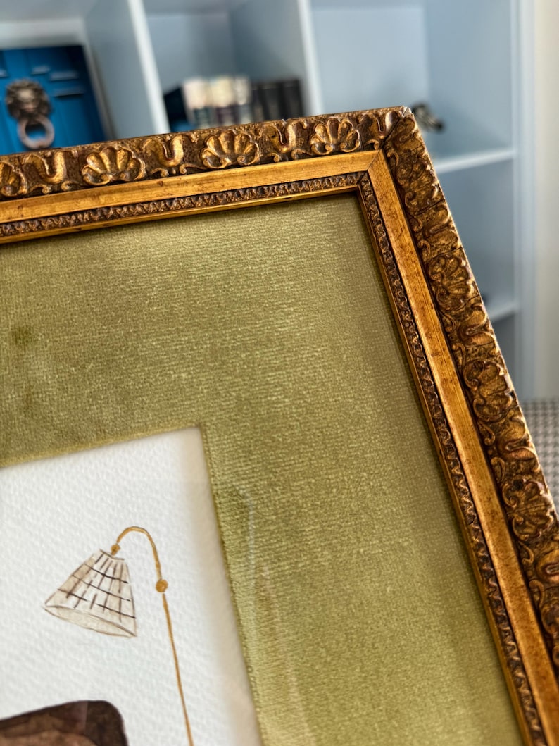 May include: A rectangular picture frame with an ornate, textured gold finish and a wide, olive green velvet mat. Inside the frame, a white paper surface features a watercolor illustration of a floor lamp with a grid-patterned lampshade.