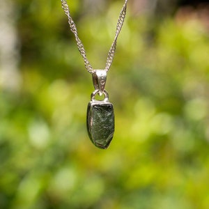 May include: A silver chain necklace with a pendant featuring a raw, green gemstone. The pendant is shaped like a teardrop and has a silver bail.