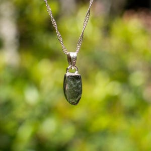May include: A silver chain necklace with a pendant featuring a raw, green gemstone. The pendant is shaped like a teardrop and has a smooth, polished surface.