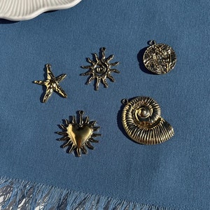 Könnte beinhalten: Eine Sammlung goldfarbener Metall-Anhänger mit nautischen Motiven, darunter ein Seestern, eine Sonne, ein Herz, eine Muschel und ein runder Anhänger mit einem detaillierten Design. Die Anhänger sind auf einem blauen Stoffhintergrund angeordnet.