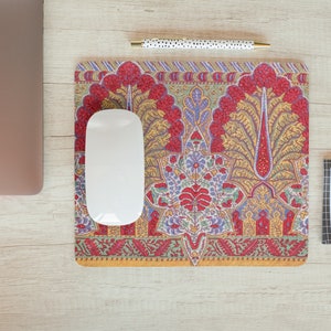 May include: A decorative mousepad with a colorful, intricate floral design in red, yellow, blue, and green. A white computer mouse sits on the mousepad. A gold binder clip, a pen, and a laptop are also visible.