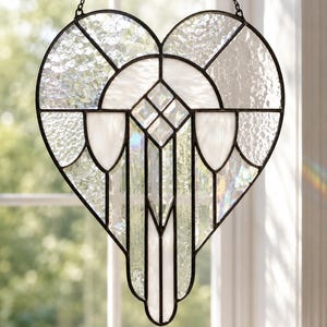 May include: A heart-shaped stained glass suncatcher featuring clear textured glass panels and opaque white glass accents held together by dark metal cames. The piece is suspended by a thin black chain and displays geometric patterns throughout.