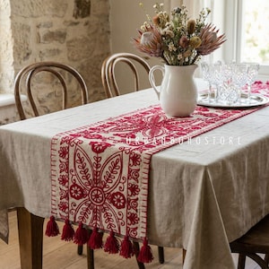 May include: A beige tablecloth with a red embroidered table runner featuring floral patterns and tassels. A white pitcher with flowers and glasses are on the table. The text "URBANBOHOSTORE" is visible on the runner.