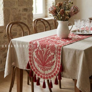 May include: A dining table set with a beige tablecloth and a red embroidered table runner. The runner features floral and leaf patterns with red tassels. A white pitcher with flowers and glasses are on the table. The text "URBAN BOHO STORE" is visible.