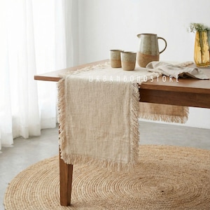 May include: A beige linen table runner with fringed edges, displayed on a wooden table. The table is set with ceramic cups, a pitcher, and a vase with flowers. A round jute rug is beneath the table. The text "URBAN BOHO STORE" is visible.