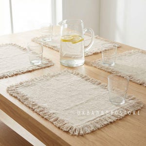 May include: A wooden table setting featuring beige placemats with fringed edges, clear drinking glasses, and a glass pitcher filled with water and lemon slices. The text "URBANBOHOSTORE" is visible on the table.