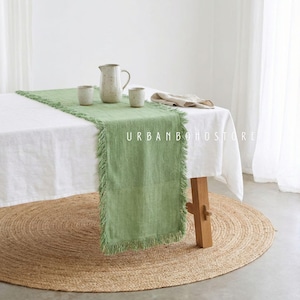 May include: A green fringed table runner is centered on a white tablecloth. A beige pitcher and two small cups sit on the runner. The text "URBAN BOHOSTORE" is printed on the runner. A round jute rug is on the floor.