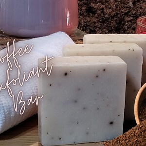 May include: Three bars of white coffee exfoliating soap with brown coffee grounds visible in the soap. The soap is sitting on a wooden surface with a small wooden bowl filled with coffee grounds. The text "Coffee Exfoliant Bar" is written in white on a white towel.