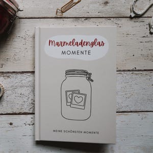 以下が含まれることがあります： 「Marmeladenglas Momente」というタイトルと「Meine schönsten Momente」というテキストが表紙に印刷された白いジャーナル。表紙には、蓋と2枚の写真が入ったガラス瓶の白黒イラストが描かれている。