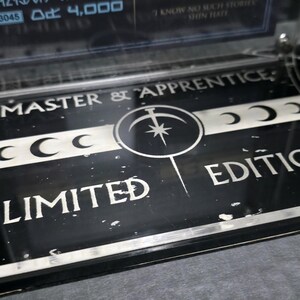 May include: Black and silver limited edition display base with the text "MASTER & APPRENTICE" and "LIMITED EDITION". The base features a central circular design with a star and crescent moons. The display is made of clear acrylic.