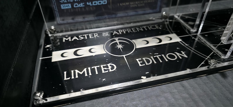 May include: Black and silver limited edition display base with the text "MASTER & APPRENTICE" and "LIMITED EDITION". The base features a central circular design with a star and crescent moons. The display is made of clear acrylic.
