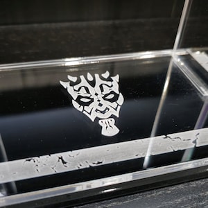 Puede incluir: Una vitrina de acr&iacute;lico transparente con un dise&ntilde;o grabado en blanco de la m&aacute;scara de un personaje de Star Wars en la parte inferior.