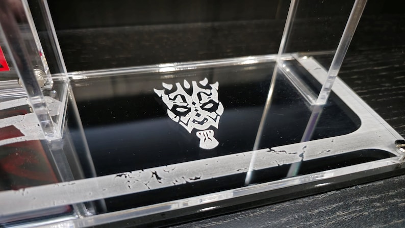 Puede incluir: Una vitrina de acr&iacute;lico transparente con un dise&ntilde;o grabado en blanco de la m&aacute;scara de un personaje de Star Wars en la parte inferior.