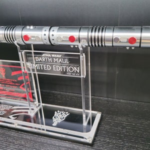 Puede incluir: Un sable de luz plateado y negro con detalles rojos en un soporte de acr&iacute;lico transparente. El soporte tiene una placa negra con el texto "STAR WARS DARTH MAUL EDICI&Oacute;N LIMITADA QUE LA FUERZA TE ACOMPA&Ntilde;E".