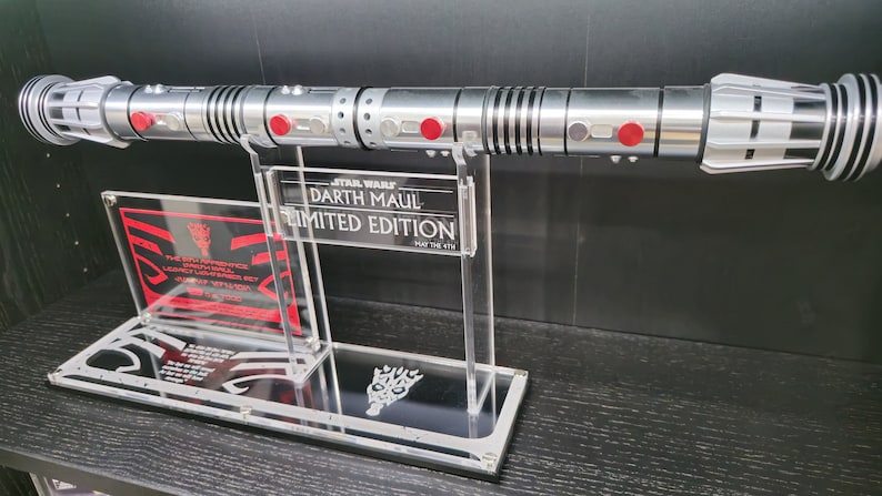 Puede incluir: Un sable de luz plateado y negro con detalles rojos en un soporte de acr&iacute;lico transparente. El soporte tiene una placa negra con el texto "STAR WARS DARTH MAUL EDICI&Oacute;N LIMITADA QUE LA FUERZA TE ACOMPA&Ntilde;E".