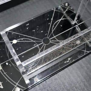 May include: Clear acrylic display stand with a black base featuring a celestial design and the text "SHINING APPRENTICE". The stand has multiple levels and is designed to hold items. The design includes white lines and dots.