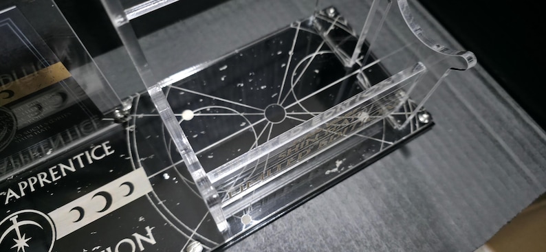 May include: Clear acrylic display stand with a black base featuring a celestial design and the text "SHINING APPRENTICE". The stand has multiple levels and is designed to hold items. The design includes white lines and dots.