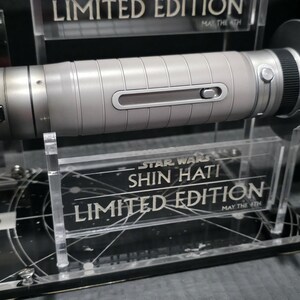 May include: A silver and black lightsaber, a limited edition Star Wars collectible, is displayed on a clear acrylic stand. The lightsaber has a detailed, metallic design with a cylindrical body and a prominent emitter. The stand features the text "SHIN HATI" and "LIMITED EDITION".