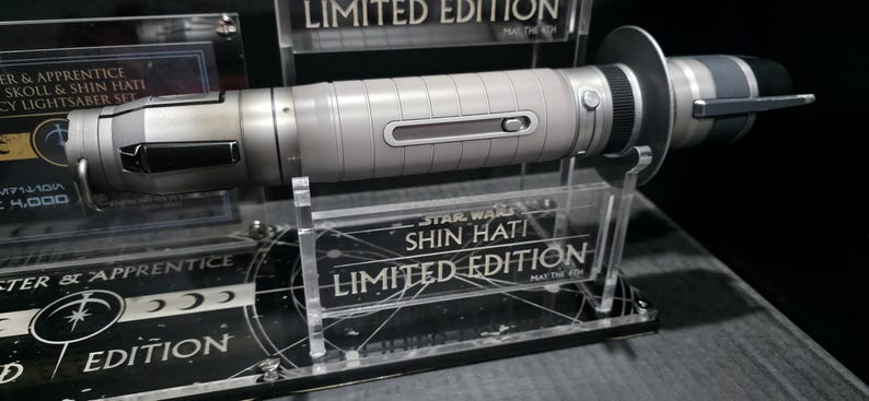 May include: A silver and black lightsaber, a limited edition Star Wars collectible, is displayed on a clear acrylic stand. The lightsaber has a detailed, metallic design with a cylindrical body and a prominent emitter. The stand features the text "SHIN HATI" and "LIMITED EDITION".