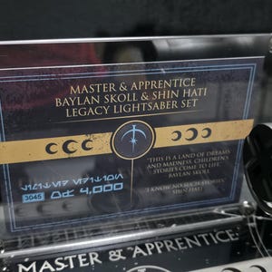 May include: A display featuring a Legacy Lightsaber Set, with text that reads "MASTER & APPRENTICE BAYLAN SKOLL & SHIN HATI LEGACY LIGHTSABER SET." The set includes a silver lightsaber and a clear acrylic sign.