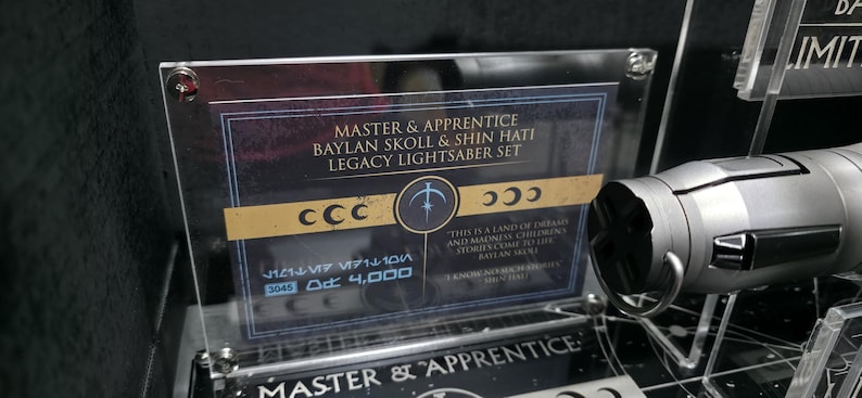 May include: A display featuring a Legacy Lightsaber Set, with text that reads "MASTER & APPRENTICE BAYLAN SKOLL & SHIN HATI LEGACY LIGHTSABER SET." The set includes a silver lightsaber and a clear acrylic sign.