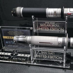 May include: A Star Wars Legacy Lightsaber Set featuring two lightsabers. The top lightsaber is black and silver, while the bottom one is silver. Both are displayed on clear acrylic stands with "LIMITED EDITION" text.