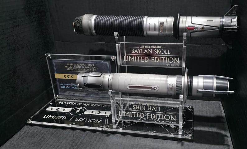May include: A Star Wars Legacy Lightsaber Set featuring two lightsabers. The top lightsaber is black and silver, while the bottom one is silver. Both are displayed on clear acrylic stands with "LIMITED EDITION" text.