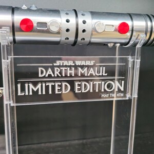 Puede incluir: Un sable de luz plateado con botones rojos y un mango negro y plateado. El sable de luz est&aacute; en un soporte transparente con un cartel que dice "STAR WARS DARTH MAUL EDICI&Oacute;N LIMITADA QUE LA FUERZA TE ACOMPA&Ntilde;E".
