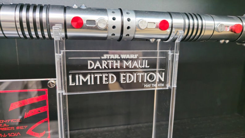 Puede incluir: Un sable de luz plateado con botones rojos y un mango negro y plateado. El sable de luz est&aacute; en un soporte transparente con un cartel que dice "STAR WARS DARTH MAUL EDICI&Oacute;N LIMITADA QUE LA FUERZA TE ACOMPA&Ntilde;E".