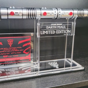 Puede incluir: Un sable de luz plateado con luces rojas en el mango, exhibido en un soporte de acr&iacute;lico transparente. El soporte tiene una placa que dice "STAR WARS DARTH MAUL LIMITED EDITION" y otra placa que dice "THE 6TH APPRENTICE DARTH MAUL LEGACY LIGHTSABER SET 1/11-V7 VF7110W 139 OUT OF 7000".
