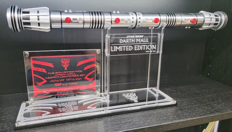 Puede incluir: Un sable de luz plateado con luces rojas en el mango, exhibido en un soporte de acr&iacute;lico transparente. El soporte tiene una placa que dice "STAR WARS DARTH MAUL LIMITED EDITION" y otra placa que dice "THE 6TH APPRENTICE DARTH MAUL LEGACY LIGHTSABER SET 1/11-V7 VF7110W 139 OUT OF 7000".