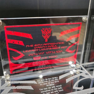 Puede incluir: Una vitrina de acr&iacute;lico transparente con una tarjeta roja y negra en su interior. La tarjeta presenta el texto "The Sith Apprentice: Darth Maul Legacy Lightsaber Set" y un gr&aacute;fico rojo de la m&aacute;scara de Darth Maul. La vitrina tambi&eacute;n tiene una peque&ntilde;a placa de acr&iacute;lico transparente con el texto "DDART LIMITED" en ella.
