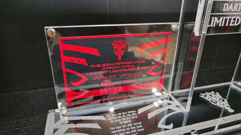 Puede incluir: Una vitrina de acr&iacute;lico transparente con una tarjeta roja y negra en su interior. La tarjeta presenta el texto "The Sith Apprentice: Darth Maul Legacy Lightsaber Set" y un gr&aacute;fico rojo de la m&aacute;scara de Darth Maul. La vitrina tambi&eacute;n tiene una peque&ntilde;a placa de acr&iacute;lico transparente con el texto "DDART LIMITED" en ella.
