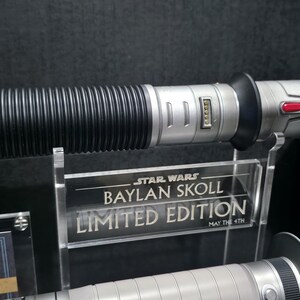 May include: A Star Wars Baylan Skoll Limited Edition lightsaber. The hilt is silver with black ribbed sections and a textured end. The display stand reads "Baylan Skoll Limited Edition".