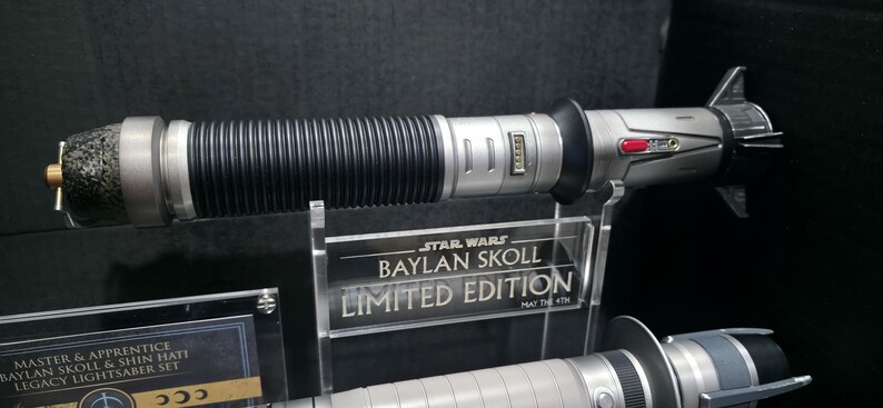 May include: A Star Wars Baylan Skoll Limited Edition lightsaber. The hilt is silver with black ribbed sections and a textured end. The display stand reads "Baylan Skoll Limited Edition".