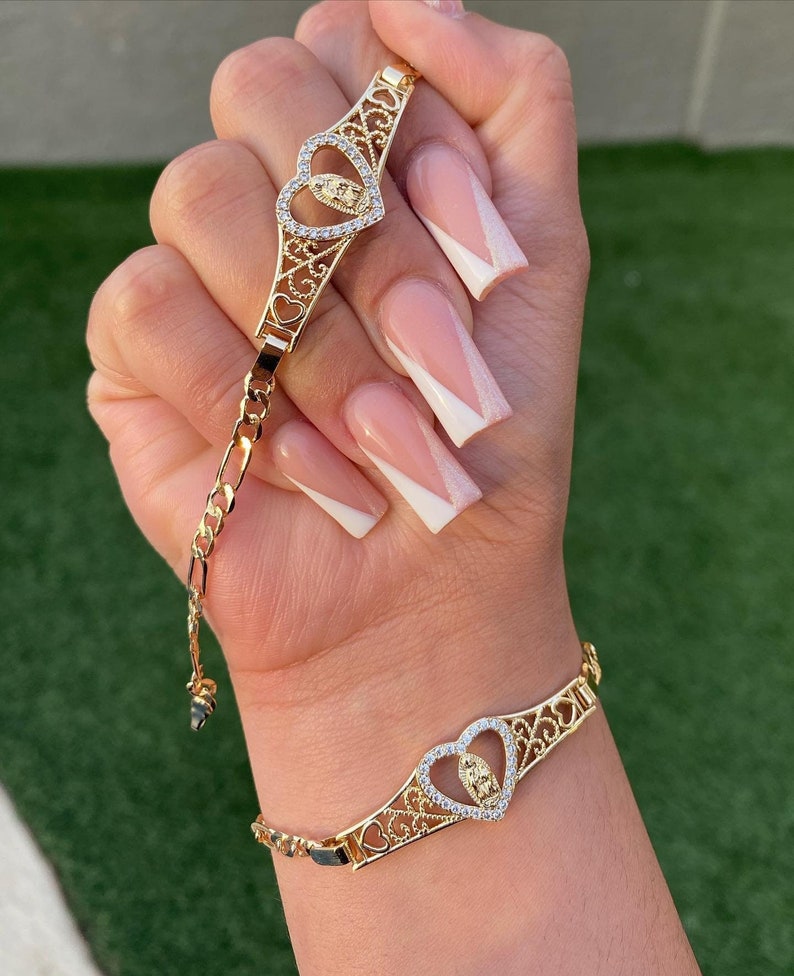 May include: Gold bracelet with a heart-shaped charm featuring a detailed image of the Virgin Mary. The charm is adorned with sparkling cubic zirconia stones.